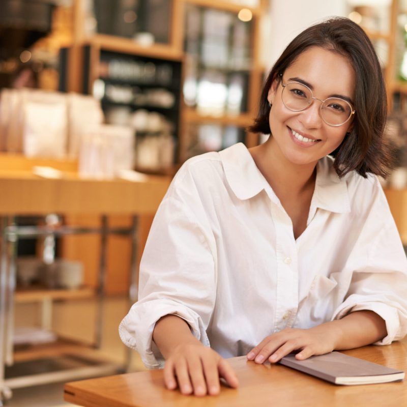 Portrait of brunette woman in glasses, smiling with confident look, sitting in cafe with notebook, working from restaurant Portrait of brunette woman in glasses, smiling with confident look, sitting in cafe with notebook, working from restaurant.
